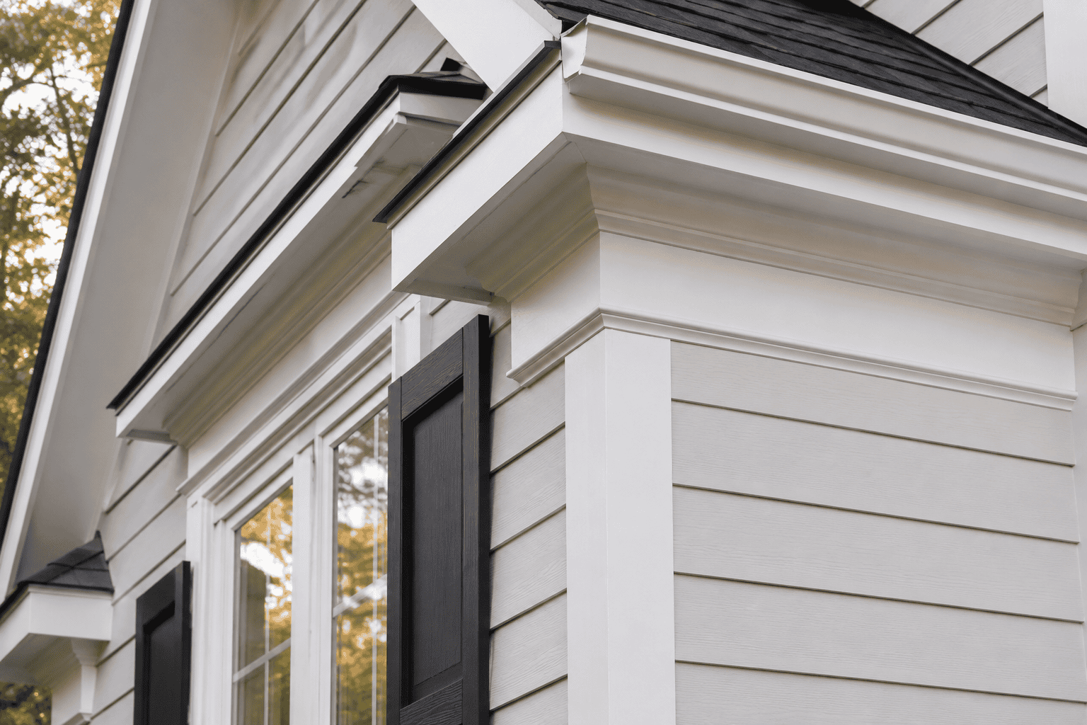 Close-up of home exterior trim detail featuring crisp white lap siding, decorative crown molding, black shutters, and dark architectural roof.