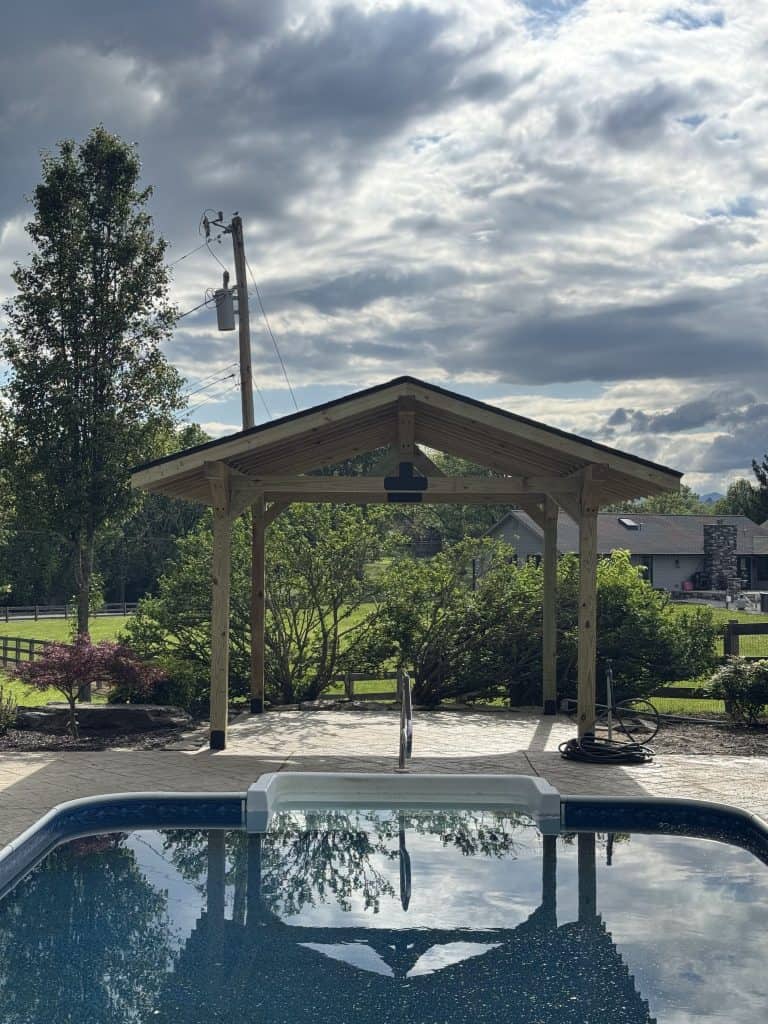 Custom timber frame pavilion reflected in pool water on stamped concrete patio with East Tennessee farmland and mountain views in background