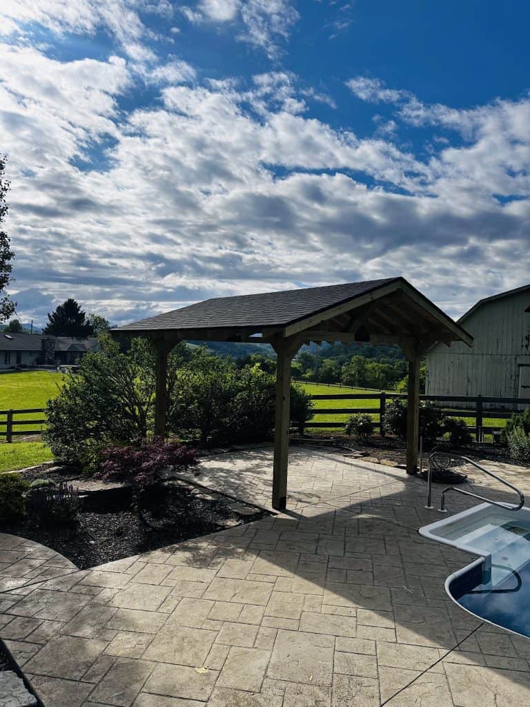 Custom timber frame pavilion with shingled roof beside a pool and stamped concrete patio in East Tennessee with mountain and farmland views