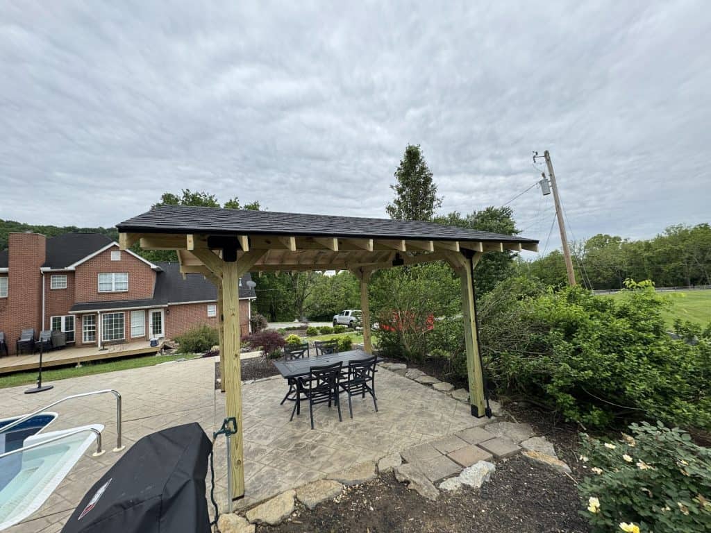 Custom timber frame pavilion with shingled roof and outdoor dining set on a stamped concrete patio beside a pool and brick home in East Tennessee