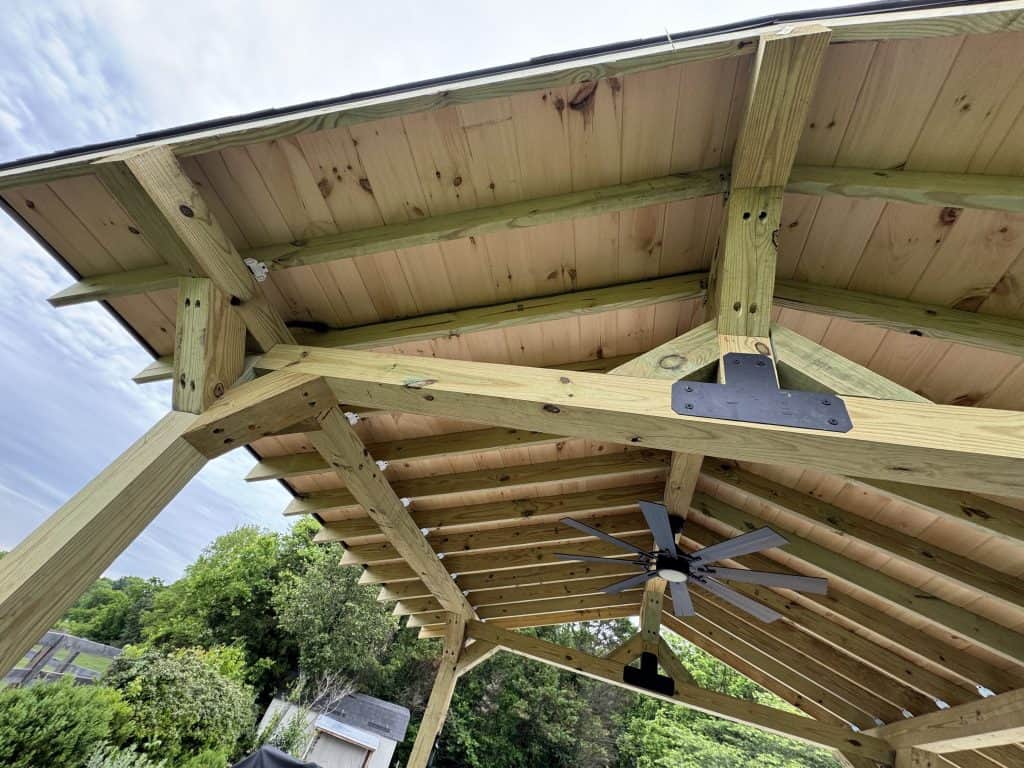 Underside view of custom timber frame pavilion showing tongue and groove ceiling boards, heavy beam framing, metal post brackets, and ceiling fan installation in East Tennessee