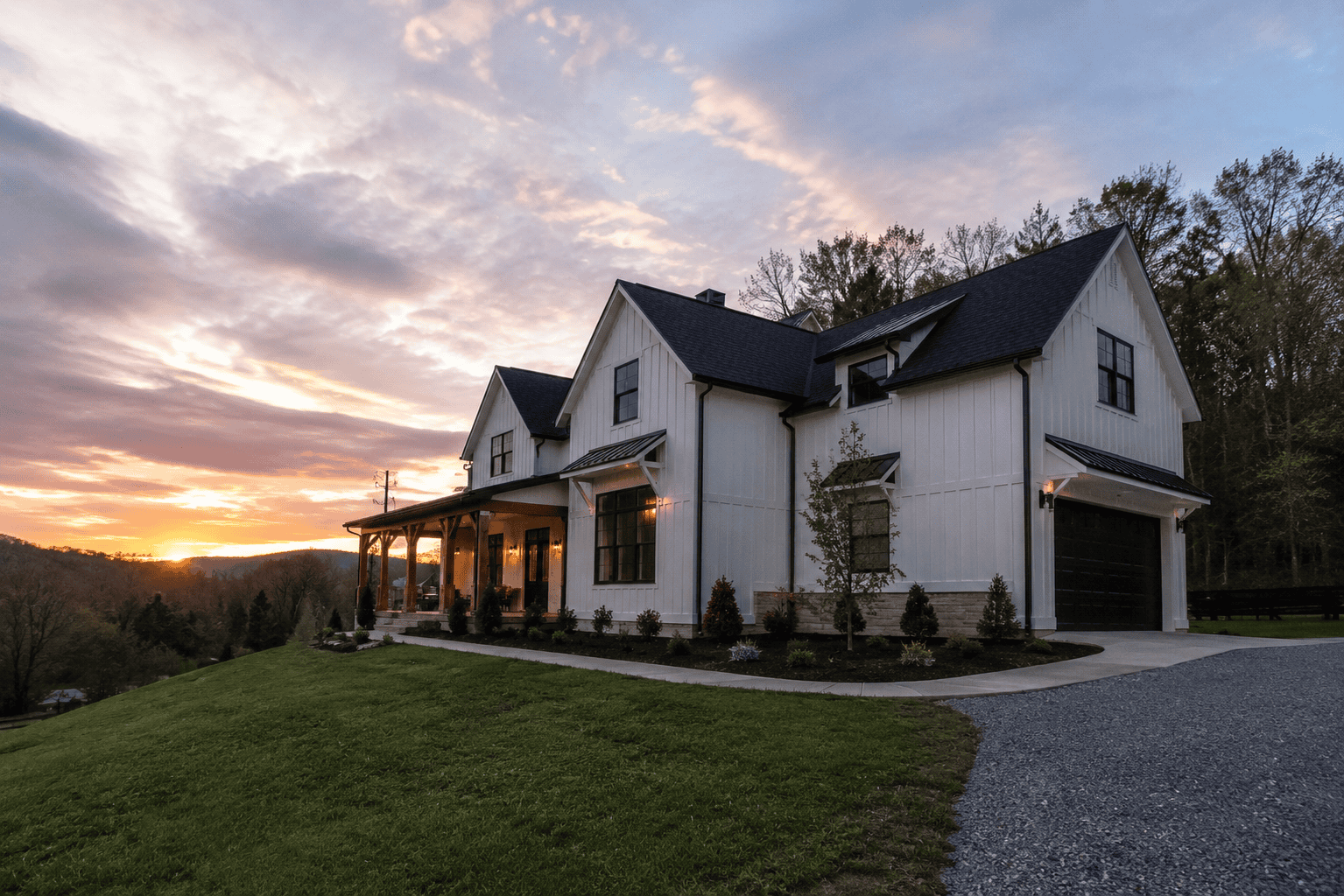 Modern farmhouse exterior at sunset with white board and batten siding, black roof, covered porch, and landscaped front yard.