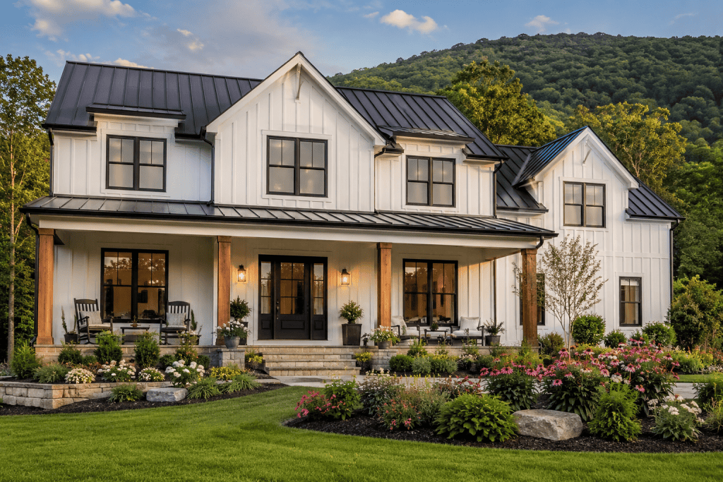 Two story white board and batten farmhouse in East Tennessee with black windows, wood porch posts, landscaped front yard with flowers and greenery, and mountain backdrop at sunset.